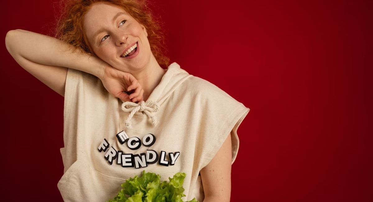 Cheerful woman with red hair in eco-friendly attire holding fresh lettuce against a vibrant red background.