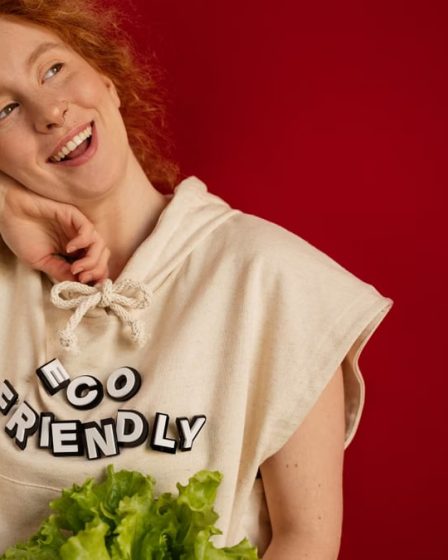 Cheerful woman with red hair in eco-friendly attire holding fresh lettuce against a vibrant red background.