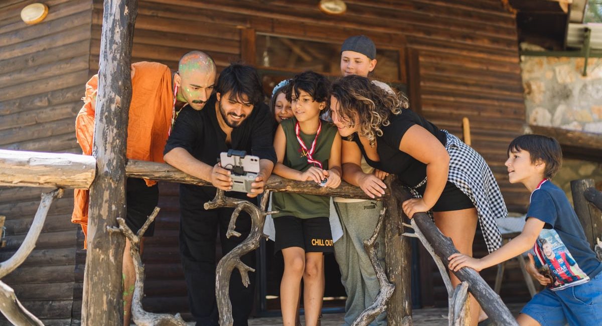 A diverse group of people joyfully watching drone footage on a wooden cabin porch.