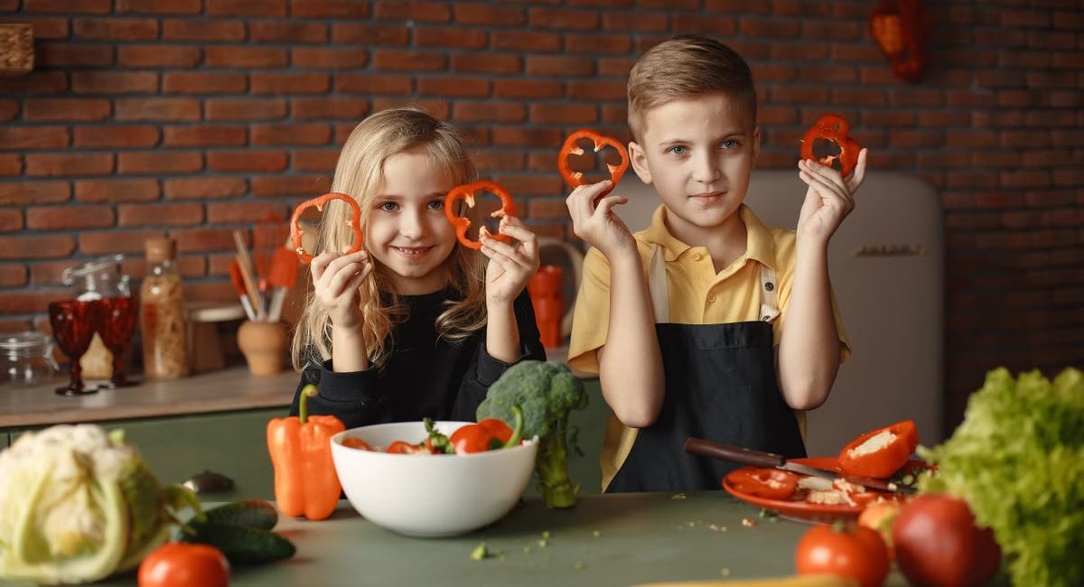 Two children holding capsicum slices while preparing vegetables in a cozy kitchen setting.