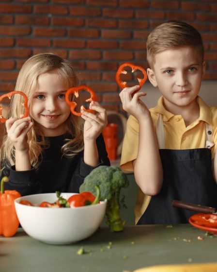 Two children holding capsicum slices while preparing vegetables in a cozy kitchen setting.
